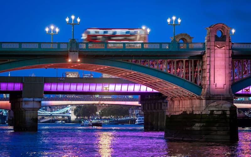 A image of a blurry red double-decker buse as it travels on a bridge over the River Thames at night.